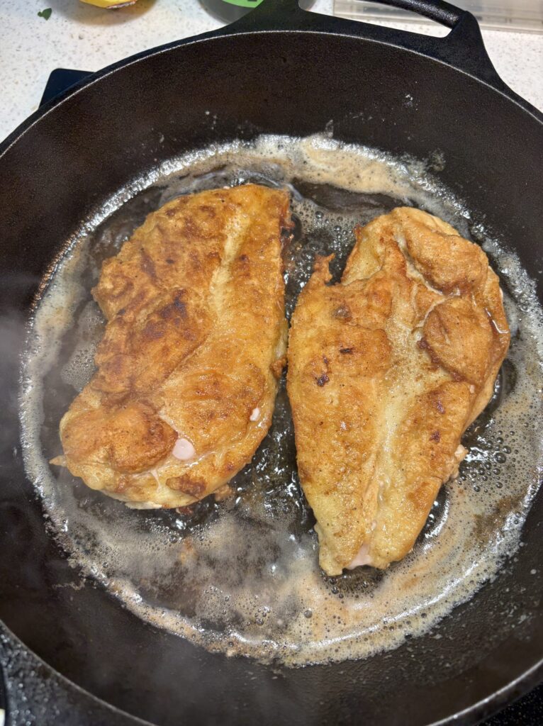 Two pieces of breaded chicken frying in a hot skillet with bubbling oil/butter around them