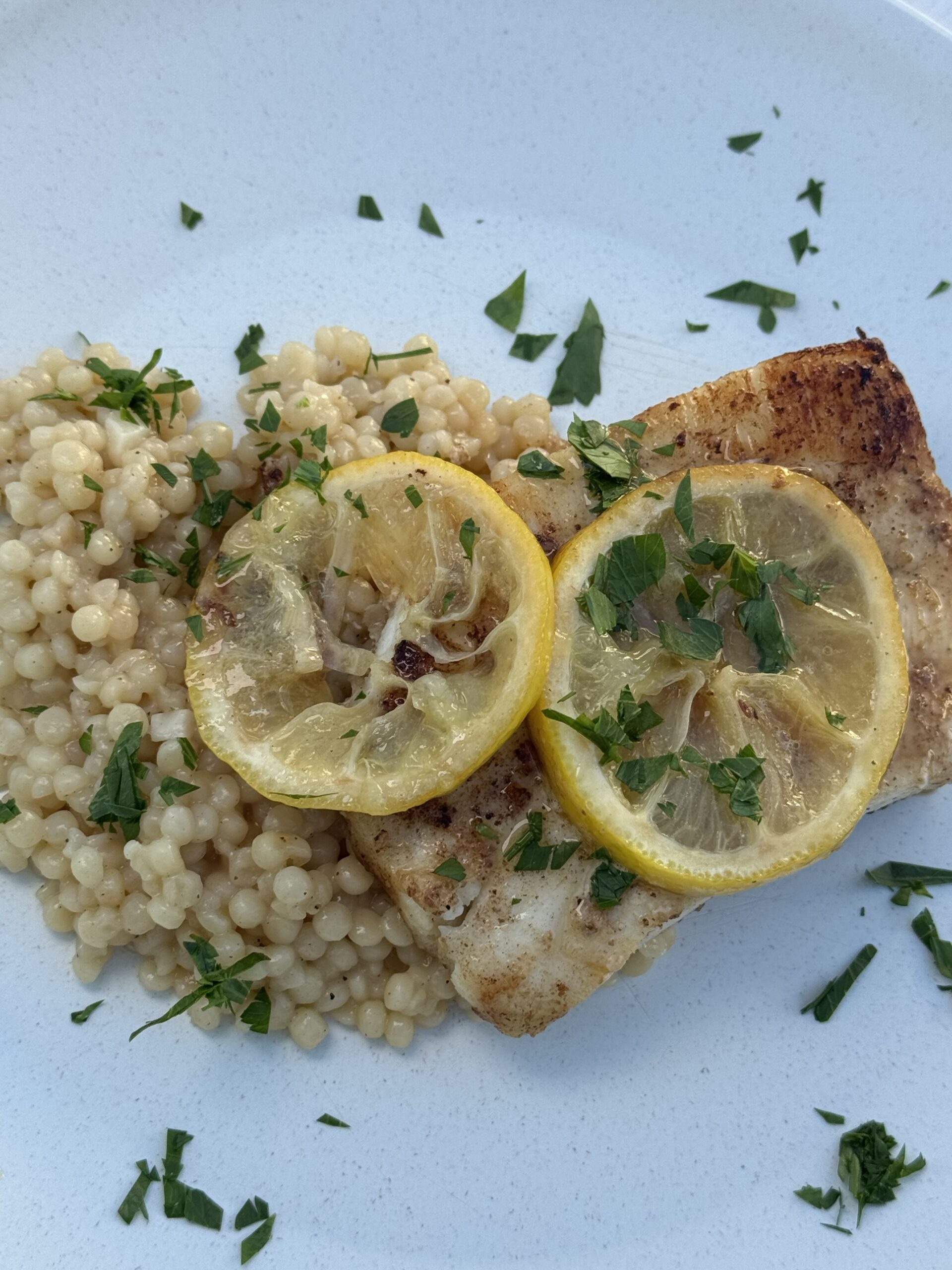 Pan-seared fish with lemon slices and parsley served over couscous on a light blue plate.